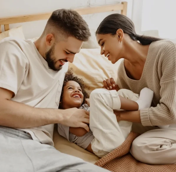 TRI_Projekt_01 Junge Familie lacht gemeinsam auf dem Bett – liebevoller Moment zu dritt.
